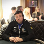 Ein glücklicher Mann sitzt an einem Pokertisch, hält zwei Karten in der Hand und lächelt selbstbewusst. In der Mitte des Tisches liegen Pokerchips und Karten. Im Hintergrund