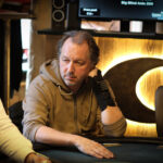 Ein Mann sitzt an einem Pokertisch, hält Karten in der Hand und betrachtet sie konzentriert. Im Hintergrund sind Pokerchips und andere Spieler zu sehen.