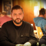 Ein Mann mit einem Pokerchip-Stapel vor sich sitzt an einem Pokertisch und betrachtet seine Karten. Andere Spieler und Dealer sind im Hintergrund zu sehen.