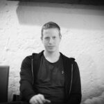 Ein Mann in einem eleganten Anzug sitzt an einem Pokertisch und hält seine Karten in der Hand, während er konzentriert auf die Mitte des Tisches schaut. Im Hintergrund sind andere Spieler und Poker