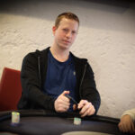 Ein Mann in einem dunkelblauen Anzug und einer roten Krawatte sitzt an einem Pokertisch und hält seine Karten in der Hand. Er hat einen konzentrierten Blick und ein selbstbewusstes L
