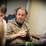 Ein Mann und eine Frau sitzen an einem Pokertisch und spielen Poker. Der Mann hält Karten in der Hand und schaut konzentriert auf den Tisch, während die Frau ihm gegenüber mit ernstem Blick ihre Chips betr