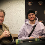 Ein Mann in einem schwarzen Anzug und einer schwarzen Sonnenbrille sitzt an einem Pokertisch und hält Karten in der Hand, während er konzentriert auf seine Gegner schaut.