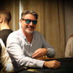 Ein Mann in einem Anzug spielt Poker an einem eleganten Tisch in einem stilvollen Casino. Er hält Karten in der Hand und schaut konzentriert auf den Tisch. Andere Spieler sind im Hintergrund