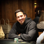 Ein Mann sitzt an einem Pokertisch mit grünem Filz und hält Karten in der Hand. Im Hintergrund sind Chips und andere Spieler zu sehen.