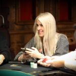Ein Mann sitzt an einem Pokertisch und hält zwei Spielkarten in der Hand. Er hat eine ernste Miene und trägt einen dunklen Anzug. Im Hintergrund sind Casino-Chips und weitere Spieler zu sehen.