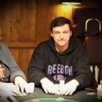 Ein Mann sitzt an einem Pokertisch und hält Karten in der Hand, während er konzentriert auf seine Gegner blickt. Im Hintergrund sind Pokerchips und Getränke zu sehen.