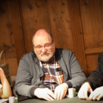 Ein Mann sitzt an einem Pokertisch und hält Karten in der Hand. Er hat eine konzentrierte Miene und trägt einen Anzug. Im Hintergrund sind weitere Spieler zu sehen.