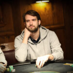 Ein Mann in einem schwarzen Anzug und einer schwarzen Sonnenbrille spielt Poker an einem Tisch, umgeben von anderen Spielern. Er hält Karten in der Hand und schaut konzentriert auf den Tisch