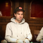 Ein Mann sitzt an einem Pokertisch und hält seine Karten in der Hand. Er schaut konzentriert auf den Tisch, um seine nächsten Züge zu planen. Im Hintergrund sind weitere Spieler und Poker