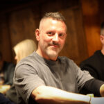 Ein Mann in einem schwarzen Anzug und einer schwarzen Sonnenbrille spielt Poker an einem Tisch. Er hält Karten in der Hand und schaut konzentriert auf den Tisch. Um ihn herum sitzen