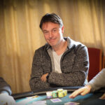 Ein Mann in einem dunkelblauen Anzug und einer Krawatte steht vor einem Pokertisch, auf dem Karten und Pokerchips liegen. Er hält zwei Karten in der Hand und schaut konzentriert auf