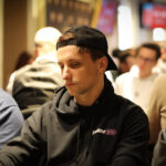 Ein professioneller Pokerspieler mit Sonnenbrille und Kappe sitzt an einem Tisch und hält eine Hand mit Spielkarten. Im Hintergrund sind weitere Spieler und Pokerchips zu sehen.