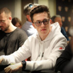Ein Mann sitzt an einem Pokertisch in einem Casino und hält zwei Karten in der Hand. Er schaut konzentriert auf den Tisch, umgeben von anderen Spielern und Pokerchips.