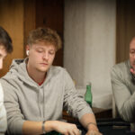 Ein Mann sitzt an einem Pokertisch und hält in seiner Hand eine Reihe von Spielkarten. Er hat einen konzentrierten Ausdruck und ist von anderen Personen umgeben, die ebenfalls Karten halten. Die Szene