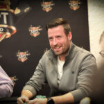 Ein Mann mit einem dunkelblauen Anzug und einer schwarzen Krawatte steht vor einem Pokertisch und hält zwei Spielkarten in der Hand. Im Hintergrund sind Pokerchips und andere Spieler zu sehen.