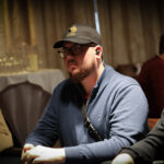 Ein Mann sitzt an einem Pokertisch mit Chips und Karten vor sich. Er hat eine ernste Miene und trägt ein dunkles Sakko. Andere Spieler sind im Hintergrund zu sehen.