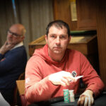Ein Mann sitzt an einem Pokertisch, der mit Chips und Karten gefüllt ist. Er betrachtet nachdenklich seine Karten, während andere Spieler im Hintergrund spielen. Die Atmosphäre ist konzent