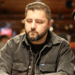 Ein Mann in einem eleganten Anzug und einer Fliege sitzt an einem Pokertisch und hält zwei Spielkarten in der Hand, während er konzentriert auf seine Gegner blickt.
