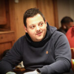 Ein Mann sitzt an einem Pokertisch und betrachtet nachdenklich seine Karten, während er von anderen Spielern umgeben ist.
