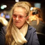 Ein Mann in einem dunkelblauen Anzug spielt Poker an einem eleganten Tisch im Casino, umgeben von anderen Spielern und einer Dealerin.