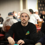 Ein Mann in einem schwarzen Anzug und mit einer Sonnenbrille sitzt an einem Pokertisch und betrachtet seine Karten konzentriert. Um ihn herum sind Pokerchips und Spielkarten verstreut.