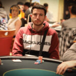 Ein Mann spielt Poker an einem Tisch mit grünem Filz. Er hat einen ernsten Gesichtsausdruck und hält Karten in der Hand. Im Hintergrund sind Pokerchips und Getränke zu