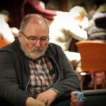 Ein Mann sitzt an einem Pokertisch und hält seine Karten mit ernstem Gesichtsausdruck. Im Hintergrund sind andere Spieler und Chips zu sehen.