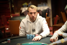 Ein Mann in einem dunklen Anzug und einer Krawatte sitzt an einem Pokertisch und hält eine Hand mit Spielkarten. Der Tisch ist mit Chips und Getränken gefüllt, im Hintergrund