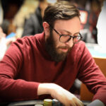 Ein Mann spielt Poker an einem Tisch in einem eleganten Casino. Er hält seine Karten in der Hand und schaut konzentriert auf den Tisch. Um ihn herum sitzen andere Spieler und beobachten das Spiel