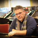 Ein Mann mit einer schwarzen Mütze und einem grauen Pullover sitzt an einem Pokertisch und hält seine Karten. Im Hintergrund sind weitere Spieler und Pokerchips zu sehen.