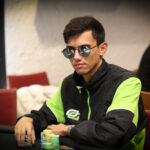 Ein Mann mit schwarzer Sonnenbrille und rotem Anzug sitzt lässig an einem Pokertisch und hält seine Karten in der Hand. Im Hintergrund sind weitere Spieler und Casino-Chips zu sehen.