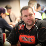 Ein Mann in einem schwarzen Anzug und schwarzen Hut sitzt an einem Pokertisch und hält seine Karten in der Hand, während er konzentriert auf das Spiel schaut.