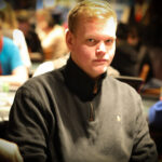 Ein Mann und eine Frau sitzen an einem Pokertisch und spielen eine Partie Poker. Der Mann hat einen Stapel Chips vor sich und hält eine Handkarte, während die Frau konzentriert ihre Karten betrachtet.
