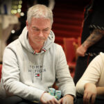 Ein Mann sitzt an einem Pokertisch und hält eine Hand mit Spielkarten. Er hat eine ernsthafte Miene und trägt ein dunkles Sakko. Andere Spieler sind im Hintergrund zu sehen, die ihre