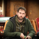 Ein Mann in einem schwarzen Anzug und einer schwarzen Sonnenbrille sitzt an einem Pokertisch und hält seine Spielkarten in der Hand. Im Hintergrund sind Casino-Chips und andere Spieler zu sehen.