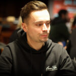 Ein Mann sitzt an einem Pokertisch und studiert konzentriert seine Karten. Um ihn herum sind Chips und Spielkarten verstreut. Im Hintergrund sind unscharfe Personen zu sehen, die ebenfalls Poker spielen