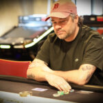 Ein Mann sitzt an einem Pokertisch und hält seine Karten in der Hand, während er konzentriert auf den Tisch schaut. Andere Spieler sind im Hintergrund zu sehen.