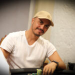 Ein Mann sitzt entspannt an einem Pokertisch und betrachtet seine Karten mit Konzentration, während im Hintergrund andere Spieler und Casino-Chips zu sehen sind.