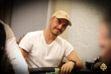 Ein Mann sitzt entspannt an einem Pokertisch und betrachtet seine Karten mit Konzentration, während im Hintergrund andere Spieler und Casino-Chips zu sehen sind.