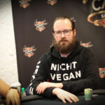 Ein Mann in einem schwarzen Anzug sitzt an einem Pokertisch und hält in der Hand ein Ass und eine Zwei in Herz. Er wirkt konzentriert und umgeben von anderen Pokerspielern in einem eleganten