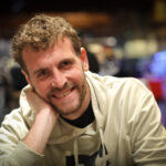 Ein Mann sitzt an einem Pokertisch und hält zwei Karten in der Hand. Er schaut konzentriert auf den Tisch, auf dem Pokerchips und Spielkarten liegen. Im Hintergrund sind weitere Spieler