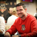 Ein Mann spielt Poker an einem Tisch mit grüner Filzdecke, umgeben von anderen Spielern. Er hält Karten in der Hand und hat einen konzentrierten Ausdruck auf seinem Gesicht.