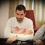 Ein professioneller Pokertisch mit grünem Filz, umgeben von Spielern, die Konzentration und Spannung ausstrahlen. Karten und Chips liegen auf dem Tisch, während im Hintergrund eine stilvolle