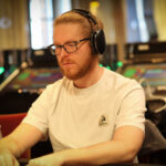 Ein Mann sitzt an einem Pokertisch mit grünem Filz und hält zwei Spielkarten in der Hand. Er hat einen konzentrierten Blick und trägt ein dunkles Hemd. Im Hintergrund sind weitere Spieler