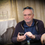 Ein Mann mit einem roten Hemd und einer schwarzen Weste sitzt an einem Pokertisch und hält seine Karten in der Hand. In der Mitte des Tisches liegen Chips und Spielkarten. Im Hintergrund