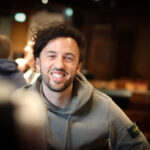 Ein Mann in einem dunkelblauen Anzug sitzt an einem Pokertisch und hält in der Hand eine Mischung aus Spielkarten und Pokerchips. Im Hintergrund sind weitere Personen und Spieltische zu sehen.