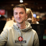 Ein Mann sitzt an einem Pokertisch und hält zwei Spielkarten in der Hand. Er schaut konzentriert auf den Tisch, umgeben von Pokerchips und anderen Spielern im Hintergrund.