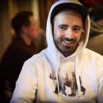 Ein Mann in einem dunkelblauen Anzug und einer weißen Hemdbluse sitzt an einem Pokertisch und betrachtet konzentriert seine Karten. Im Hintergrund sind Pokerchips und eine Glühlampe