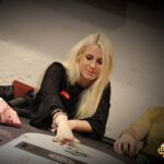 Ein Mann in einem schwarzen Anzug und einer Krawatte sitzt an einem Pokertisch und hält seine Karten in der Hand, während er konzentriert auf die Mitte des Tisches schaut. Um ihn herum